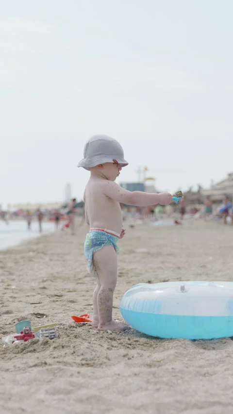 Young child wearing a sun hat plays with colorful toys on sandy beach near Vidéo 341438279