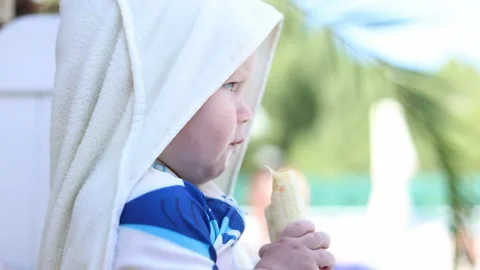 Young child wrapped in a towel eats a banana beside a bright outdoor swimming Stock Footage 347686552