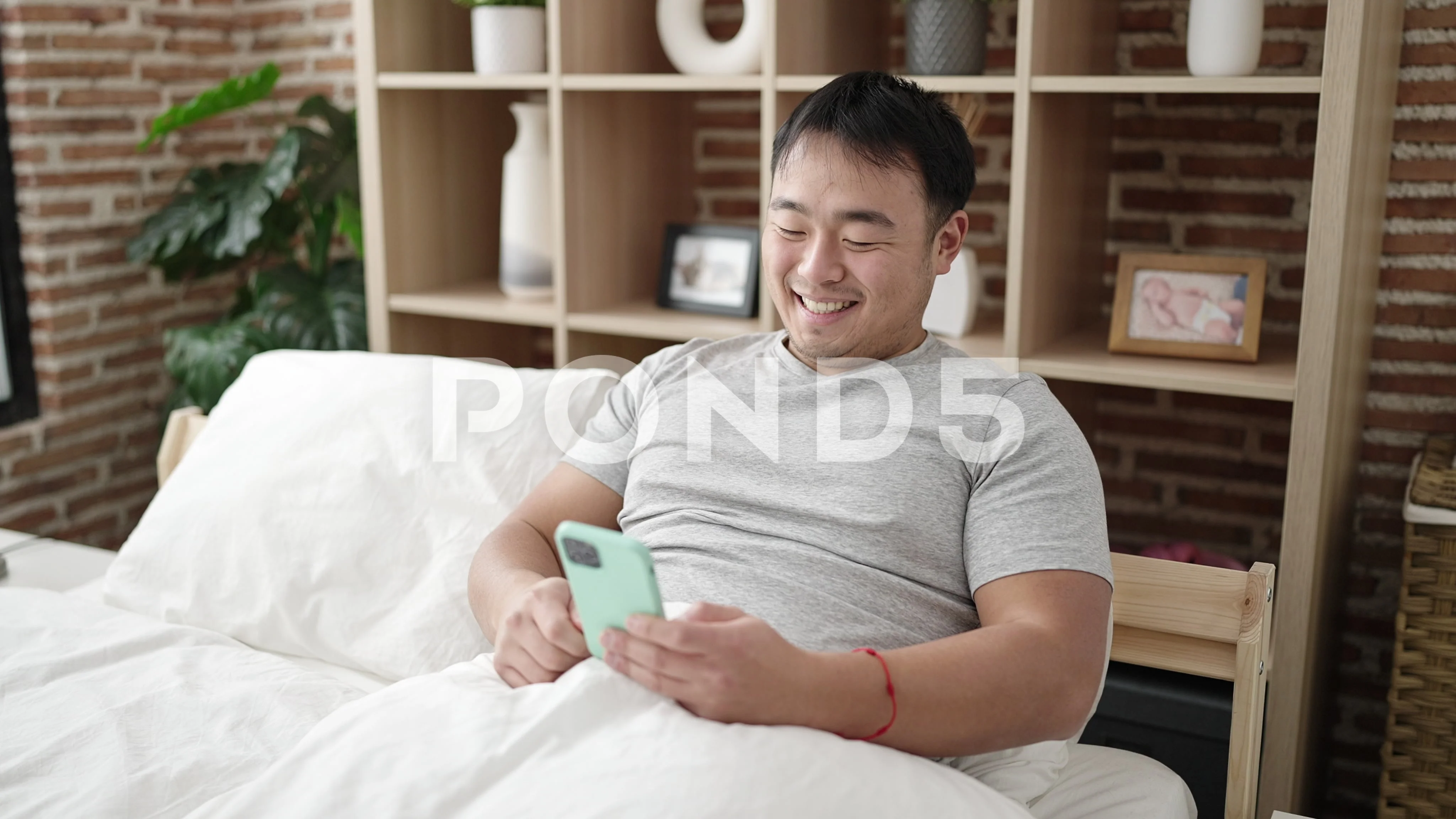 Young chinese man having video call sitting on bed at bedroom