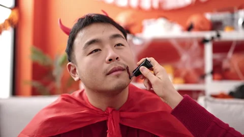 Young chinese man wearing devil costume make up looking on mirror at home Stockbeeldmateriaal 236767147