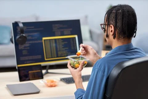 Young computer programmer eating takeout lunch at workplace while writing code Stock Photos