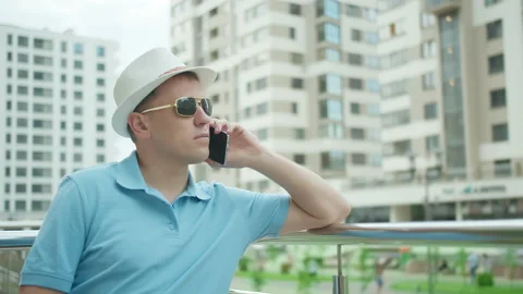 Young confident man in hat talking on mobile phone while standing near fence Stock Footage 152796482