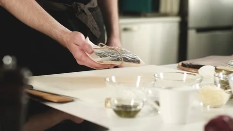 Young cook in an apron puts piece of fresh fish wrapped in parchment on table. Stock Footage 136508267