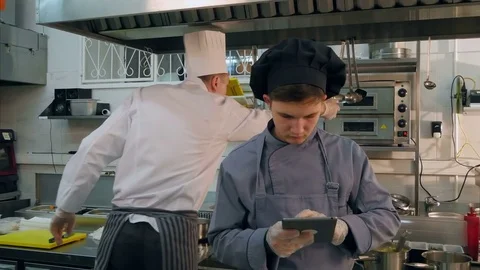 Young cook trainee using tablet while chef busy cooking in the kitchen Stock Footage 72323199