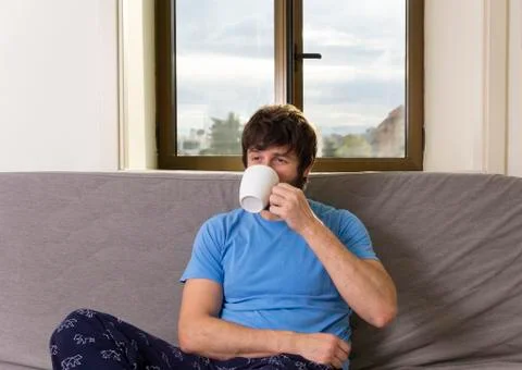 Young cool man sitting on a sofa at living room with a cup. Pensive expressio Stock Photos