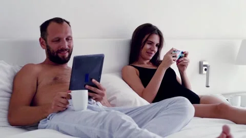 Young couple in bedroom; Man using tablet, woman playing game on smartphone 스톡 동영상 91240758