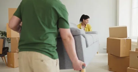 Young couple carrying gray sofa in new apartment with moving boxes around Stock Footage 340919210