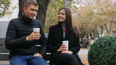 Young couple drinking coffee and talking while sitting on a bench in the street Stock Footage 97144830