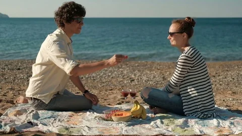 Young couple eating fruit sitting on beach on summer day. Stock Footage 77610197