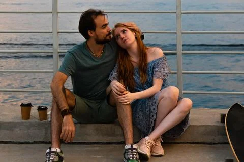 Young couple embracing while sitting on embankment against river Stock Photos