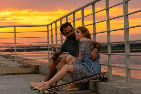 Young couple embracing while sitting on embankment against sunset sky Stock Photos