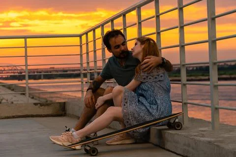 Young couple embracing while sitting on embankment against sunset sky Stock Photos