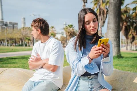 Young couple experiencing tension while one browses on a smartphone in a sunny Stock Photos