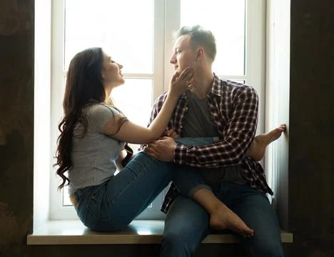 Young couple hugging while sitting on the windowsill by the window Stock Photos