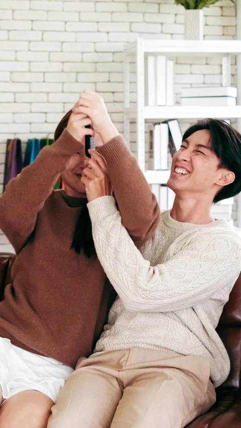 A young couple laughs and playfully fights over the TV remote on a cozy sofa Stock Footage 325708294