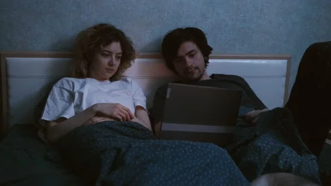 A young couple is lying in bed and watching something on a laptop Stockbeeldmateriaal 197558366