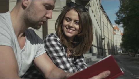 Young couple reading together while sitting in front on stairs, outdoors. 動画素材 54416397
