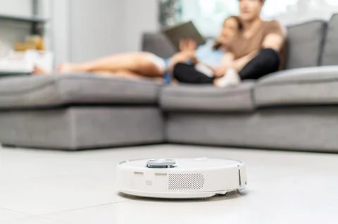 Young couple resting on the couch while robotic vacuum cleaner doing its work Stock Photos