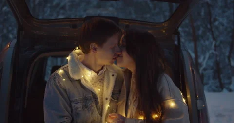 Young couple sit in a car trunk surrounded by christmas lights embracing Stock Footage 152467648