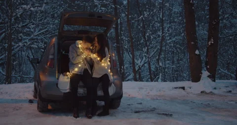 Young couple sit in a car trunk surrounded by christmas lights embracing Stock Footage 152468091