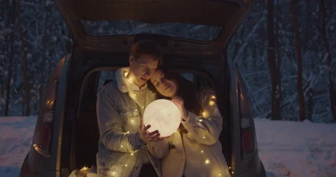 Young couple sit in a car trunk surrounded by christmas lights embracing Stock Footage 152468792