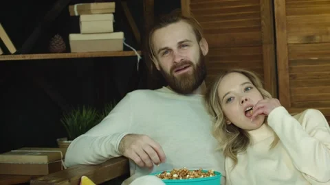 Young couple is siting on a sofa eating popcorn Watching TV at home Stock Footage 147036268