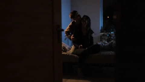 A young couple sits on a bed, hugging and kissing under warm evening lighting, s Stock Footage 296424561