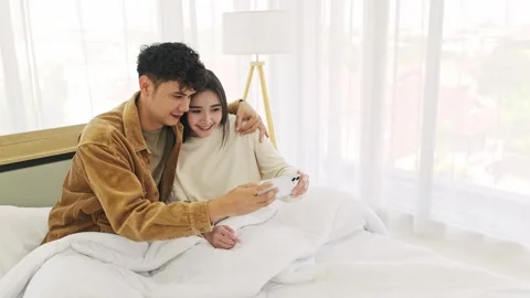 A young couple sits on a bed, smiling and looking at a smartphone together 스톡 동영상 327122782
