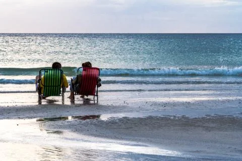 Young couple sits in chairs watching sunset as incoming tide washes over thei Stock Photos