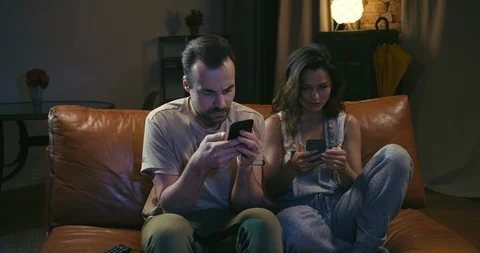 Young couple sits on couch and is distracted by TV program from texting on 스톡 동영상 108676421