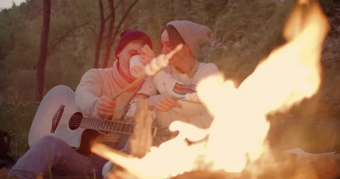 A young couple sits by the fire, talking, a guy fries marshmallows and drink tea Stock Footage 114754737