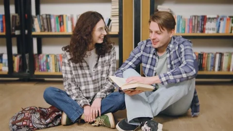 Young couple sits on the floor of a library sharing a book and discussing its Stock Footage 318094537