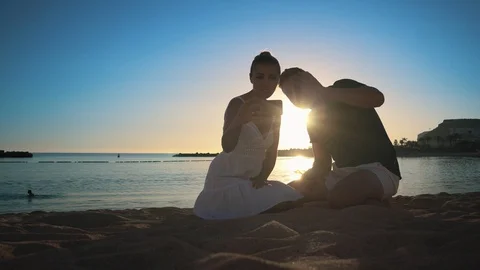 A young couple sits on a sandy beach at sunset and communicates with friends via Stock Footage 104201488