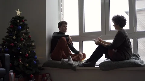 A young couple sits talking and chatting at a large window next to a Christmas  Stock Footage 118538015