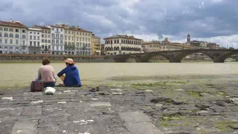 Young couple sitting on the Arno river shore, on a cloudy spring day. 库存影片 133123748