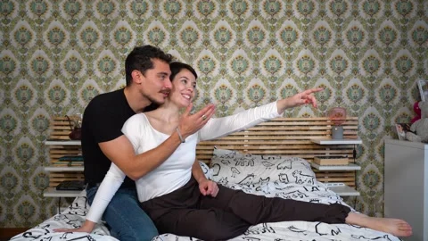 Young couple is sitting on the bed and pointing at something at the window. Stock Footage 229650040