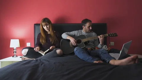 Young couple sitting on the bed reading and playing the guitar Stock Footage 129859067