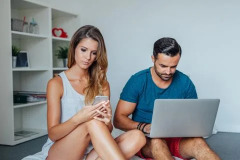 Young couple is sitting on the bed while using pc laptop and mobile phone. Stock Photos