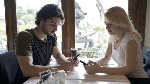 Young couple sitting at cafe table and texting messages on smartphone exchang Stock Footage 78119230
