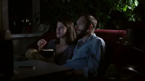 Young couple sitting on a couch in front of a TV screen and eating popcorn Video stock 118278929