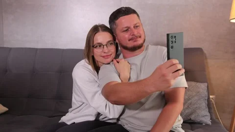 A young couple sitting on a couch, taking a selfie with a cellphone. Stock-Footage 290411614
