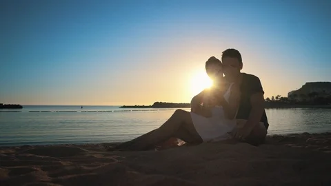 Young couple sitting embracing on a sandy beach at sunset and chatting with Stock Footage 104205074