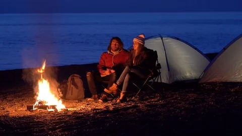Young couple sitting by the fire. camping with tents by the sea. spring evening. Stock Footage 107783579