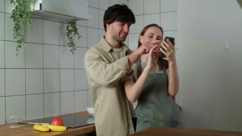 Young couple is sitting at the kitchen counter, looking at a smartphone and 库存影片 239671825