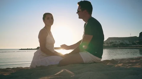 A young couple is sitting on the sandy seashore, smiling, holding hands and Stock Footage 104248971