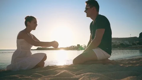 A young couple is sitting on the sandy shore of the sea, smiling and taking each Stock Footage 104247719