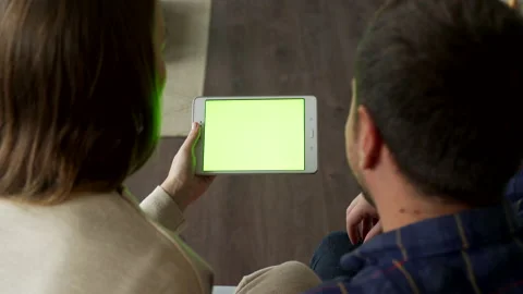 A young couple sitting on a sofa can be seen over their shoulder using a green 库存影片 144838955