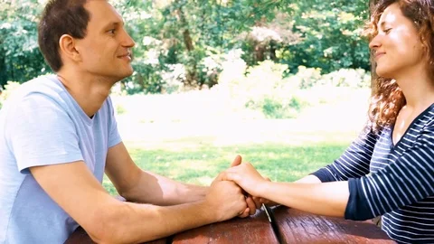 Young couple sitting, talking, holding hands at picnic table in outdoor park Stock Footage 80207615