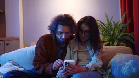 Young couple sitting together on sofa using mobile phone or smartphone, internet Stock Footage 242513521