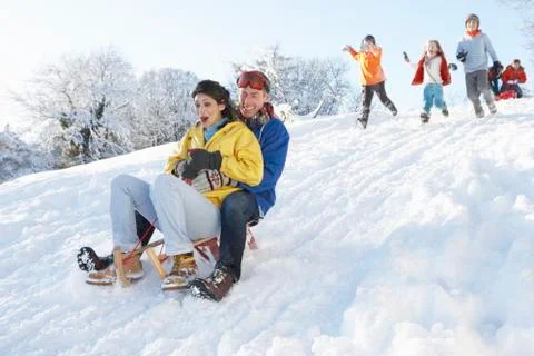 Young couple sledging down hill with family watching Stock Photos